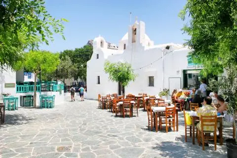 Outdoor stone plaza with wooden tables and chairs in front of a white church, people dining on the right