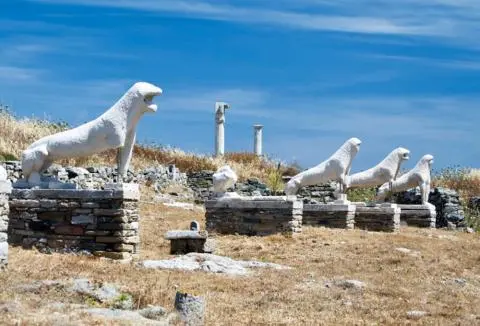 Row of white lion-like marble statues on stone plinths amid dry grass under a blue sky