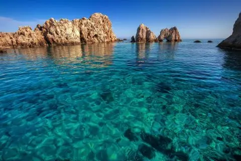 Turquoise clear sea in foreground with sunlit rocky islets and jagged rock formations