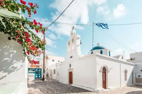 White church with bell tower, blue dome and Greek flag on a stone-paved street