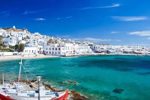 Mykonos seaside with white buildings on hillside, turquoise water and a red-white small boat in foreground
