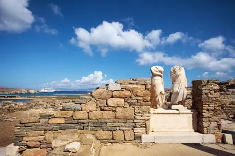 Two headless ancient marble statues on a stone wall by the Aegean shore with a cruise yacht in the distance