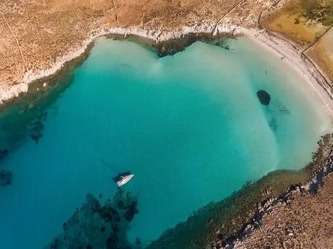 A sailboat anchored in clear turquoise water off a curved sandy coastline with rocky edges