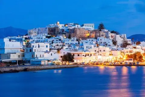 Seaside town at dusk with white buildings and lit waterfront reflecting on calm water