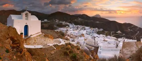 Small white chapel with blue door on hillside, white village and sunset over sea