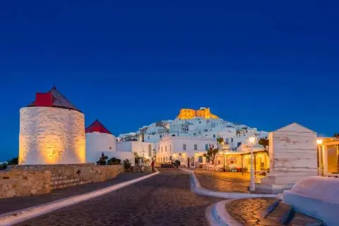 Illuminated white windmills and cobblestone promenade at dusk with lit white buildings on hill
