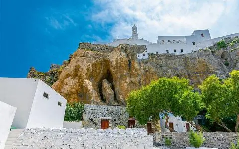 White stone buildings and cave entrance beneath a rocky cliff on a sunny Aegean coastline