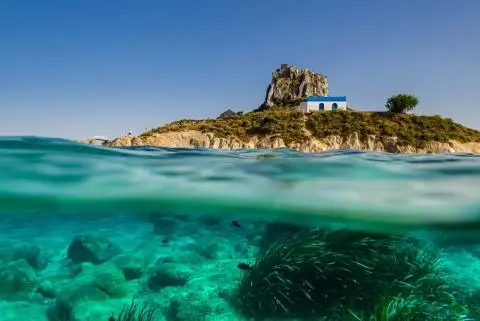 Turquoise shallow sea with underwater rocks and a white building with blue roof on a rocky hill