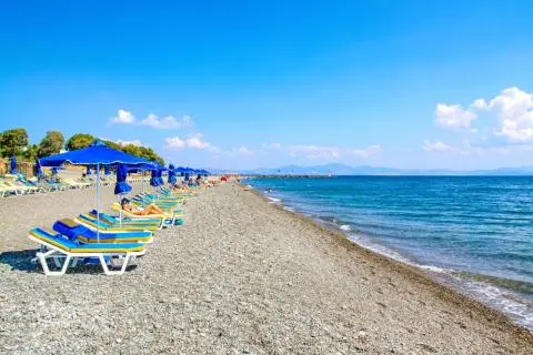 Pebble beach with rows of blue umbrellas and yellow-blue loungers, calm sea on the right