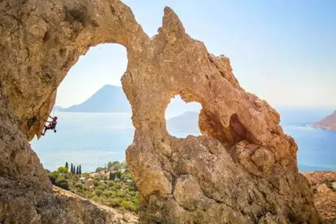 Person rock climbing on a rope beneath a natural rock arch with sea and islands visible