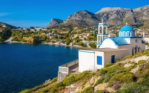 Blue-domed Greek coastal church on rocky hillside overlooking deep blue sea and village
