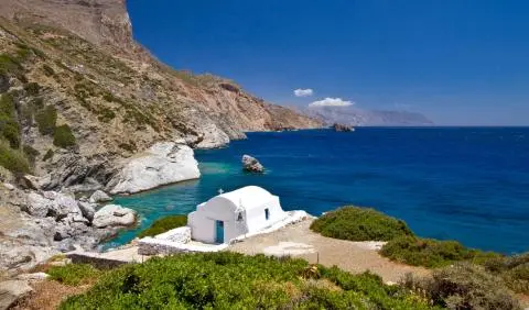 Small white seaside chapel by turquoise water, rocky coastal cliffs and deep blue sea on a sunny day