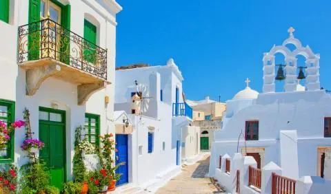 Sunlit Aegean island street with whitewashed houses, green doors, balcony and bell tower