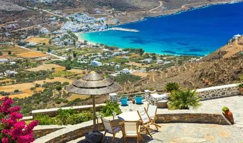 Hillside terrace with straw umbrella, table and chairs overlooking turquoise bay and white coastal village