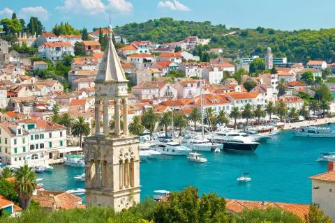 Stone bell tower in foreground with palm-lined harbor, yachts and red-tiled Dalmatian town