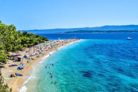 Long sandy beach lined with pine trees, clear turquoise sea and crowded sunbathers with umbrellas along shore