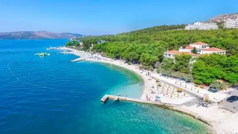 Sandy beach with yellow umbrellas and sunbeds by turquoise sea, pier and pine-covered shoreline