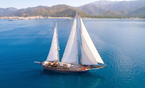 A three-masted sailing yacht with white sails on calm blue sea, mountains along the shore in the background