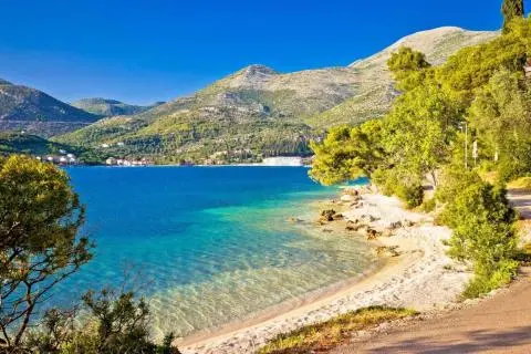 Turquoise bay with pebbled shore, pine trees on the right and mountains on horizon