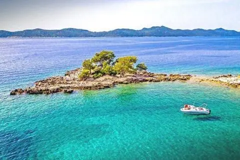Small rocky islet with trees in turquoise sea and a white boat anchored to the right