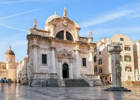 Baroque stone church façade with steps and statues on a sunlit plaza