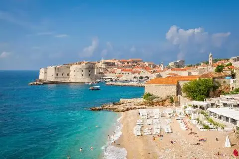 Turquoise sea and beach in front of historic city walls and red-tiled roofs of Dubrovnik harbor