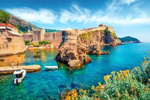 Historic stone walls and rocky cliff by turquoise Adriatic water with small boats