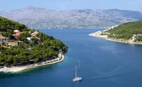 White sailing yacht cruising on deep blue water near a pine-covered cove with red-roof houses