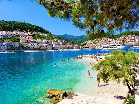 Pebble beach with swimmers in turquoise bay, coastal stone houses and a docked yacht