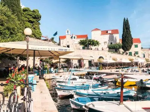 Small boats moored at a marina with waterfront restaurant tables and stone buildings
