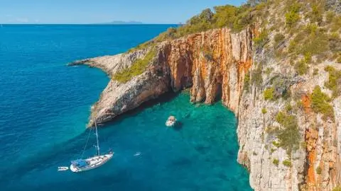 White sailing yacht and small motorboat anchored in turquoise cove beneath orange-hued rocky cliffs