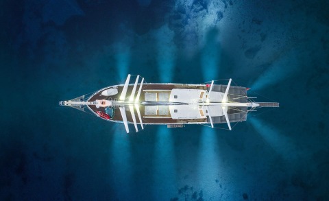 Overhead view of a large yacht with a wooden deck and white structures, bright lights on the deck, deep blue sea in the background