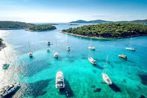 Aerial view of turquoise bay with multiple yachts and sailboats anchored near wooded islands