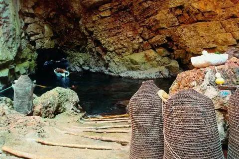 Rock-walled sea cave entrance with woven fish traps in foreground and small boat tied at water