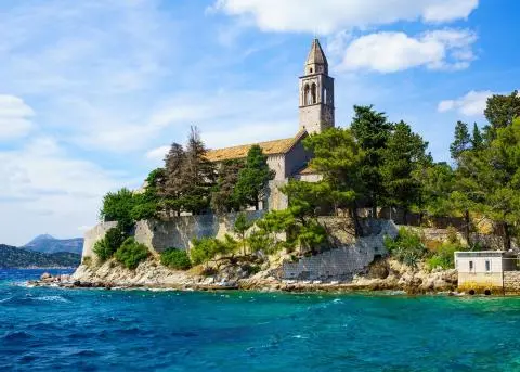Stone-walled island church with pine trees overlooking turquoise sea and rocky shore