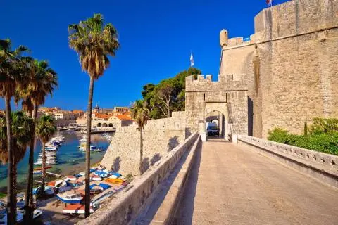 Stone bridge leading to a fortress gate with castle wall on right and palm trees and moored boats at harbor left