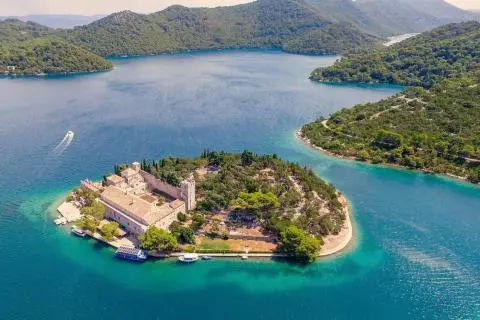 Historic stone complex on a small wooded island with docked boats, surrounded by turquoise sea
