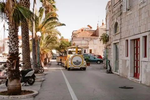 Yellow tourist train driving down a palm-lined seaside street past stone buildings