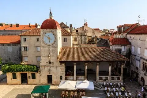 Historic stone clock tower with red-tiled roof and adjoining colonnaded building, terrace seating below