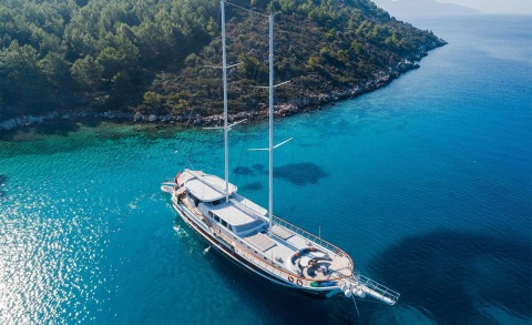 Catamaran-style sailing yacht with two masts anchored in turquoise water near a rocky shoreline