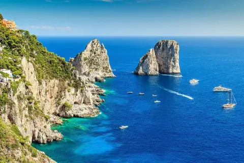 Several yachts and small boats sailing near rocky sea stacks and steep coastal cliffs in clear blue water