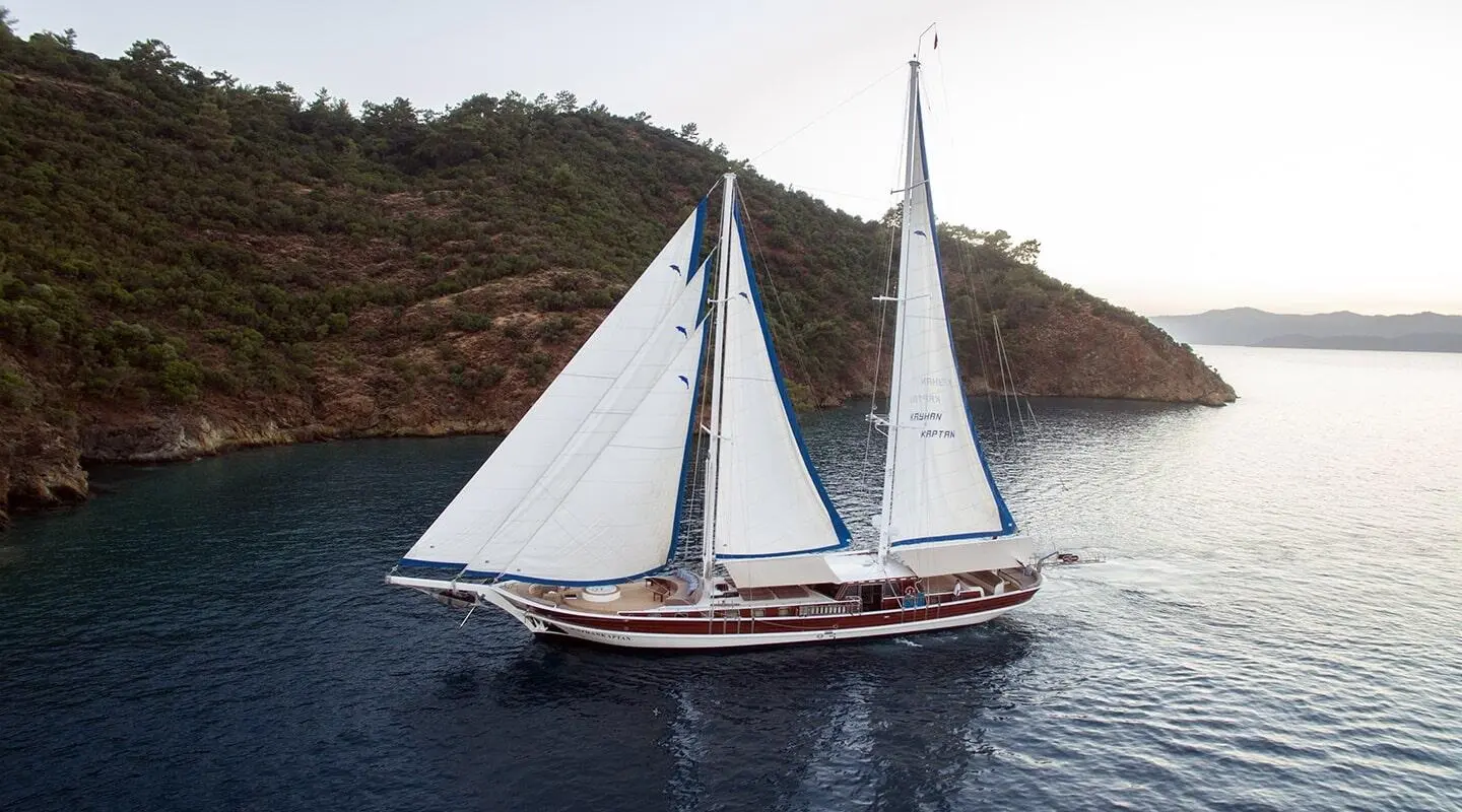 Two sails on a classic yacht gliding near rocky coastline at sunset