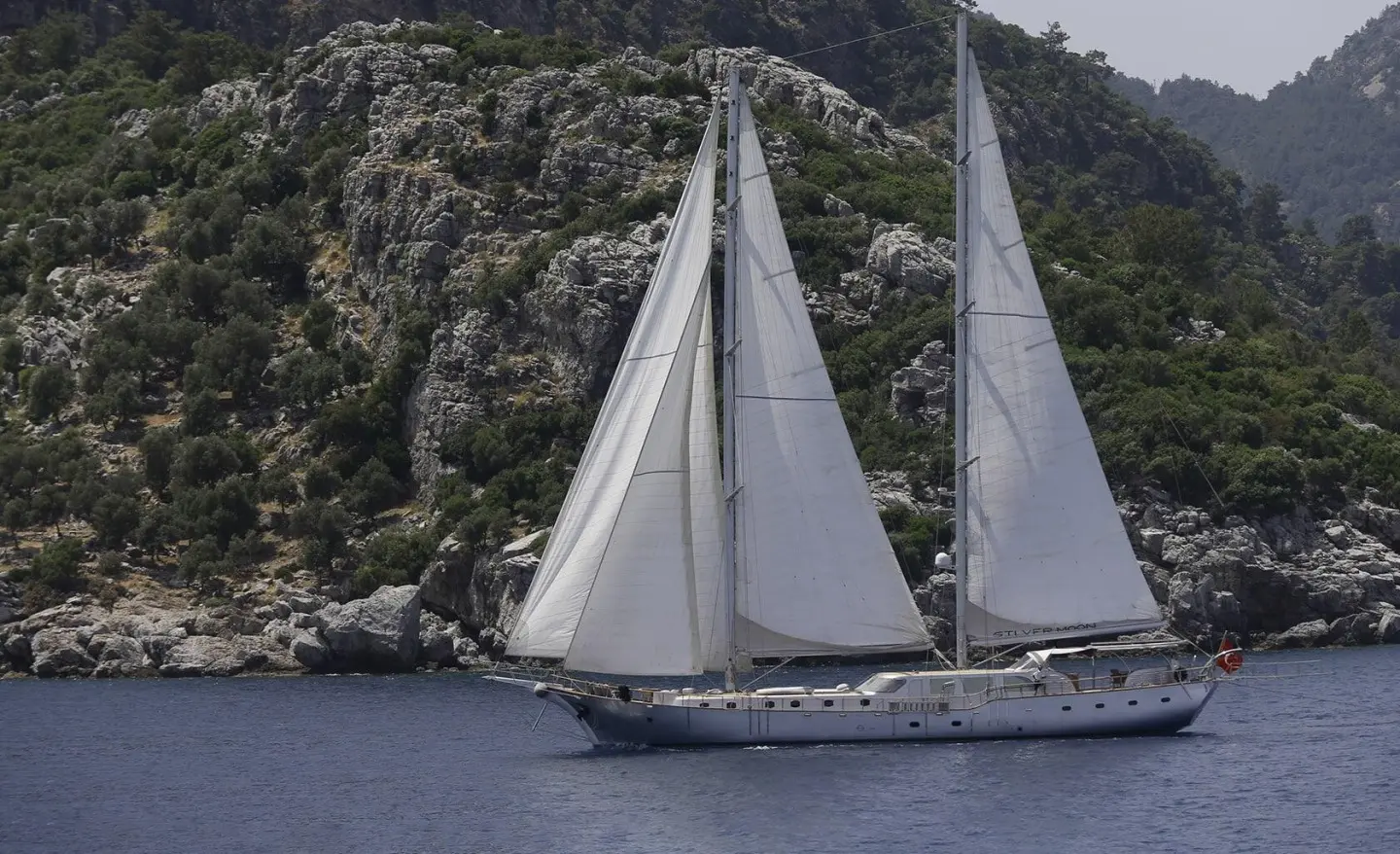Silver Moon sailing near rocky coastline with white sails unfurled