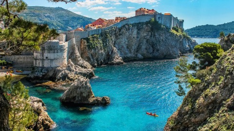 Historic stone city walls on rocky cliffs above turquoise Adriatic water with two people kayaking