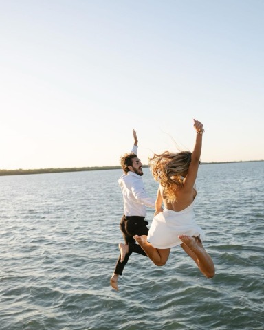 Woman in white dress and man in shirt jumping together off into the sea with visible waves