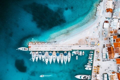 Aerial view of white sailboats and motorboats moored along a pier, turquoise sea and coastal houses