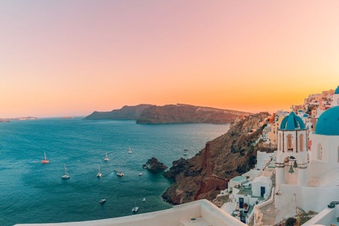 White cliffside houses and blue-domed church in Santorini, sailboats on sea at sunset