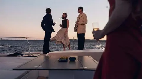Three well-dressed people standing on a yacht at sunset holding wine glasses