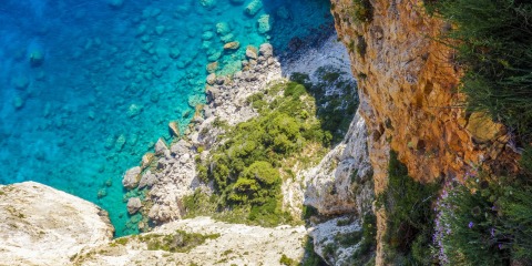 Turquoise sea seen from cliff edge with rocky shoreline, green shrubs below