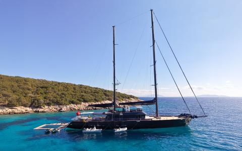 Two-masted black-hulled sailing yacht anchored in clear blue water near rocky coastline